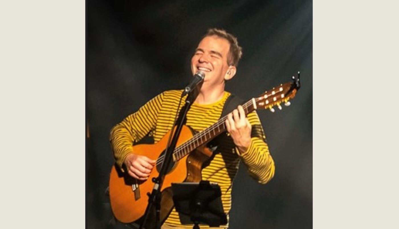 Un homme blanc portant un chandail jaune sur scène qui joue de la guitare et chante au microphone.